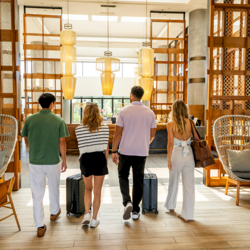 Four people entering a modern, brightly lit lobby with luggage, featuring stylish decor and wooden elements.