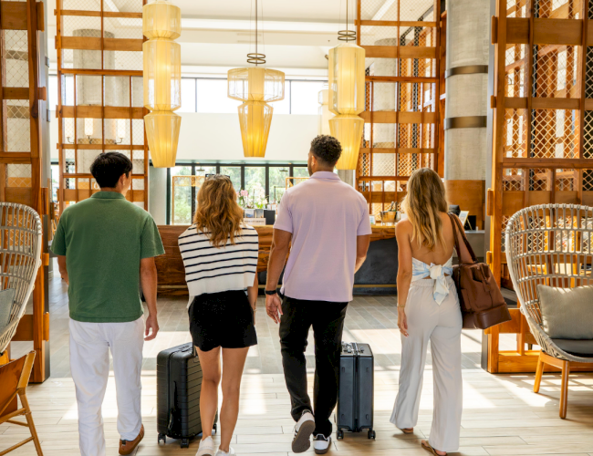 Four people entering a modern, brightly lit lobby with luggage, featuring stylish decor and wooden elements.
