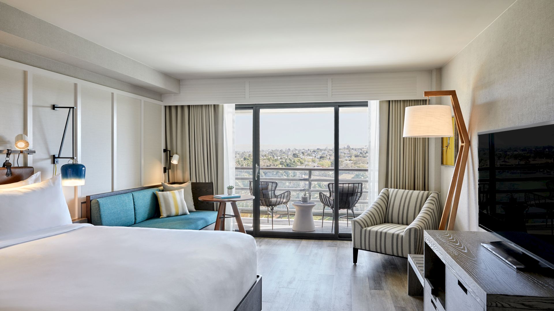 A modern hotel room with a large bed, sofa, TV, and seating, opening to a balcony with a scenic view outside.