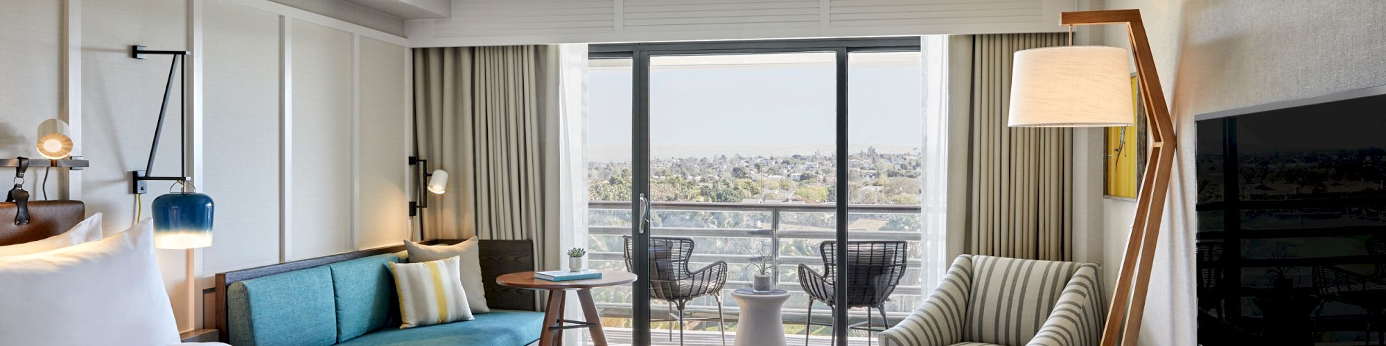 A modern hotel room with a large bed, sofa, TV, and seating, opening to a balcony with a scenic view outside.