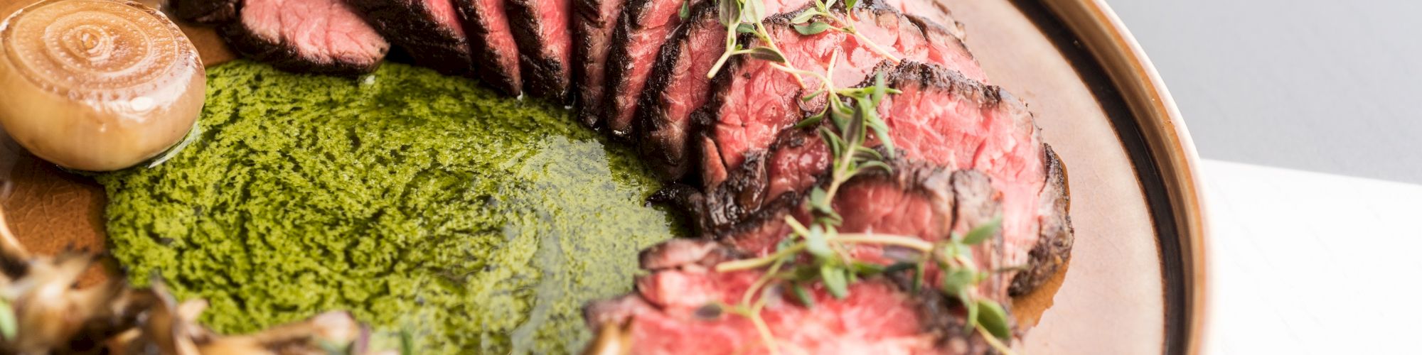 The image shows a plate with sliced steak garnished with microgreens, accompanied by a green puree, mushrooms, and an onion, presented artfully.