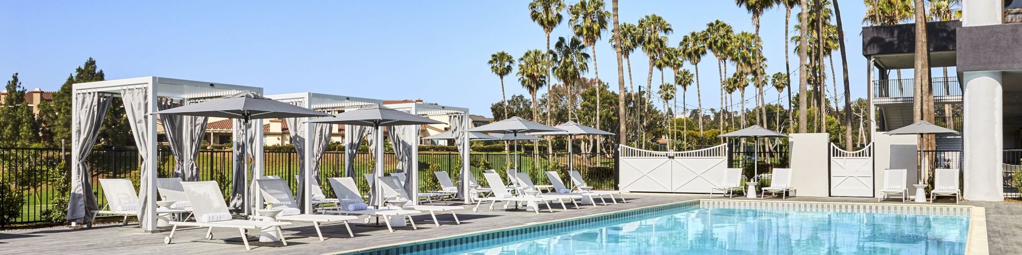 A serene pool area with lounge chairs, cabanas, and umbrellas, surrounded by palm trees and a clear blue sky.