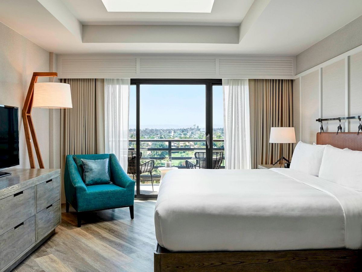 A hotel room with a bed, TV, green chair, lamps, dresser, and a balcony with a scenic view visible through large sliding glass doors.