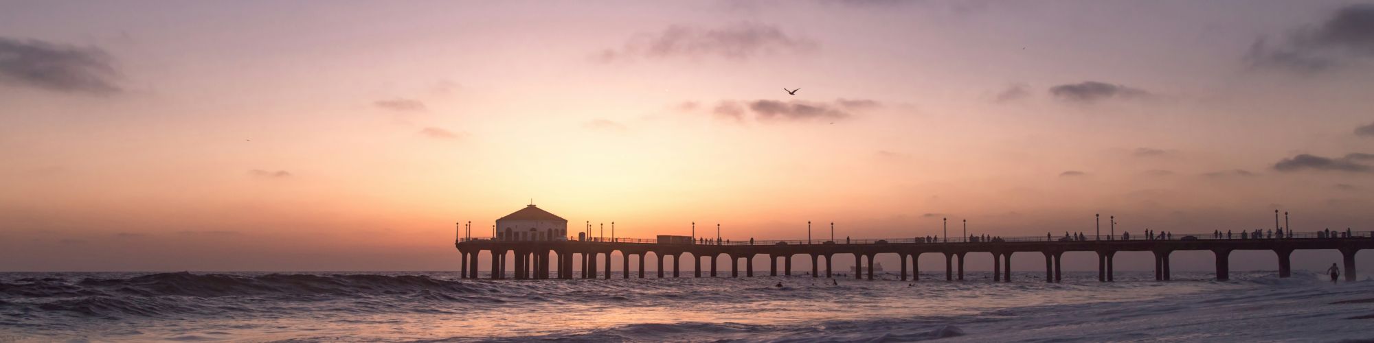 A picturesque sunset over an ocean pier, with calm waves rolling onto the shore and a bird flying in the distance.