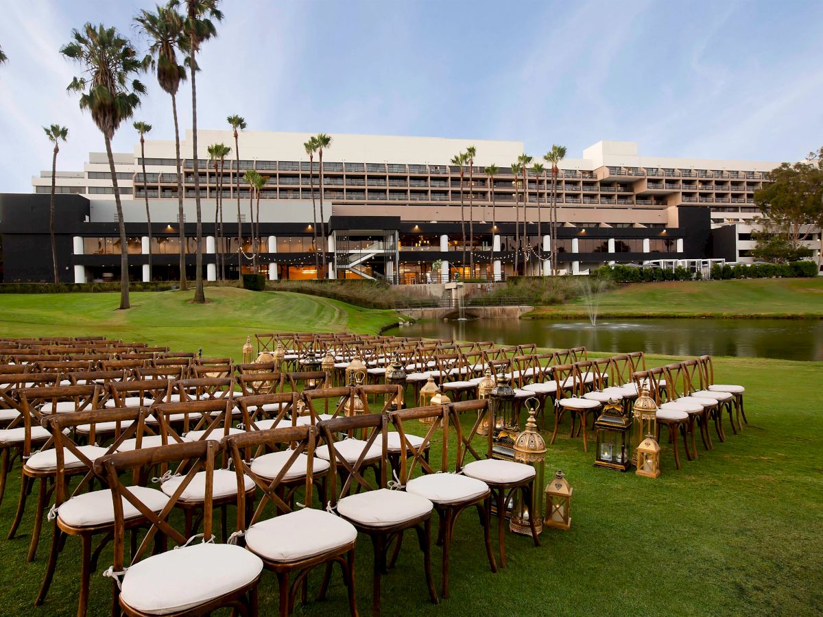 Chairs are set up outdoors facing a large building, likely for an event. Palm trees and a pond are in the background, creating a serene setting.