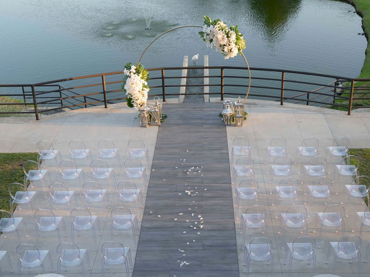 An outdoor wedding setup by a lake with rows of clear chairs, a flower-decorated arch, and a wooden aisle strewn with flower petals ends the sentence.