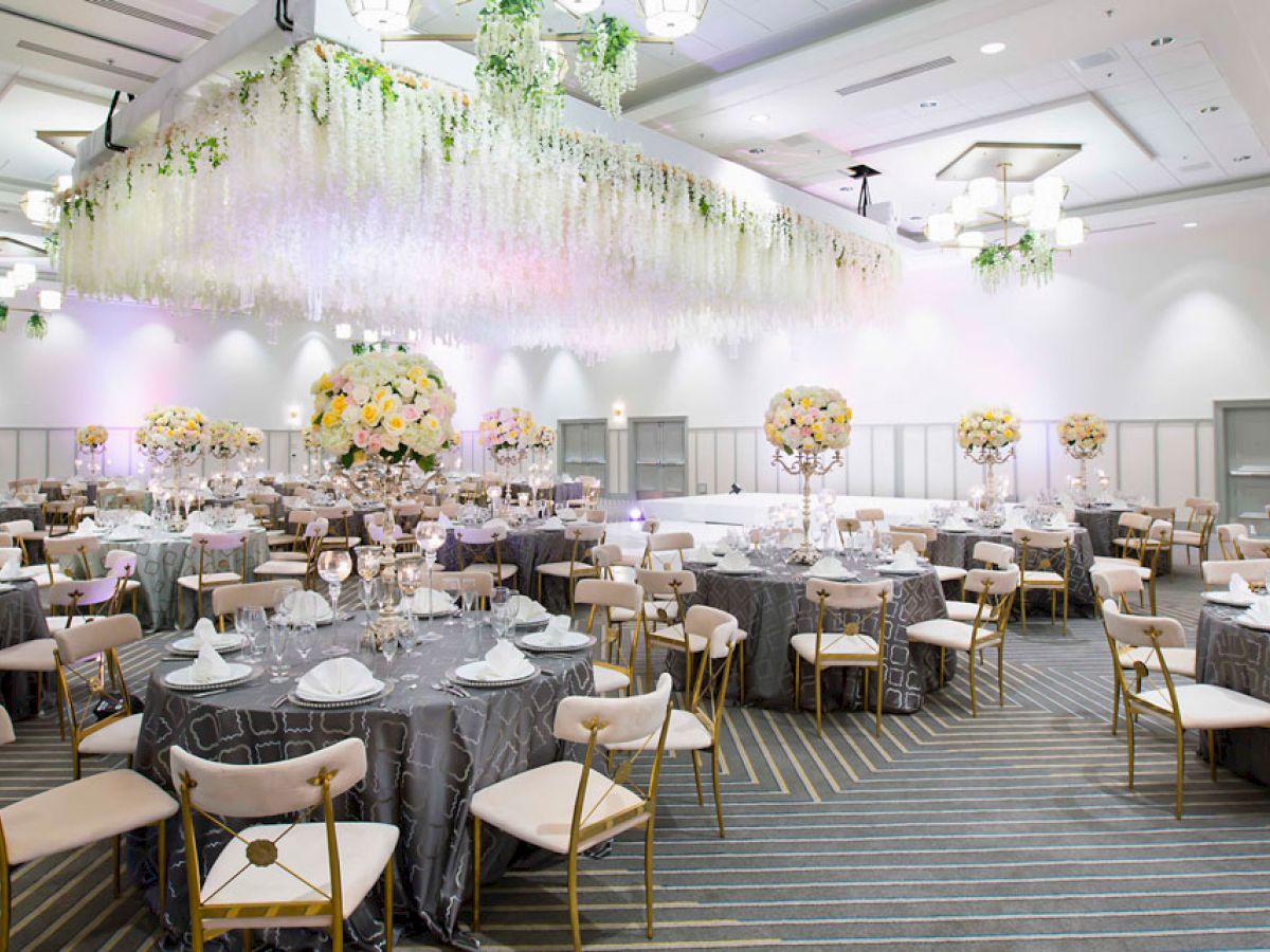 The image shows an elegantly decorated banquet hall with round tables, floral centerpieces, and a hanging floral arrangement from the ceiling.