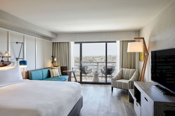 A modern hotel room with a bed, a couch, a TV, and a balcony with chairs and a table overlooking a scenic view, lit naturally.