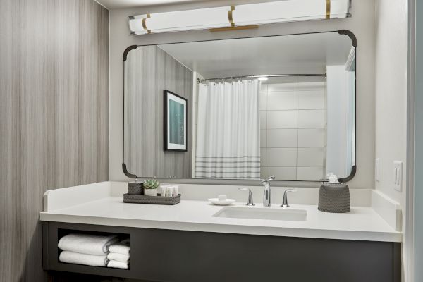 A modern bathroom with a large mirror, sink, folded towels, and toiletries on the countertop, and a shower with a white curtain in the background.