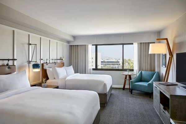 The image shows a modern hotel room with two beds, a window view, a blue armchair, a floor lamp, and a TV on a cabinet.