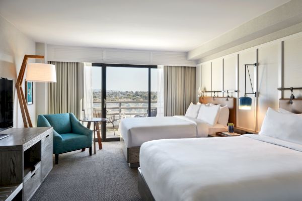 The image shows a modern hotel room with two large beds, a blue armchair, a floor lamp, a TV on a console, and a balcony with a city view.