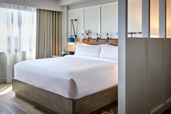 The image shows a modern hotel room with a large bed, white bedding, a wooden bed frame, a hanging lamp, and light streaming through curtains.