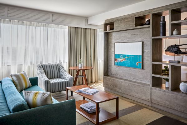 A modern living room with a sofa, striped armchair, wooden coffee table, TV, shelves, and large windows with sheer curtains.