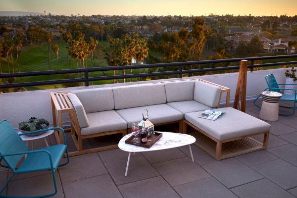 A rooftop patio with modern outdoor furniture, including a sectional sofa, table, chairs, decor, and distant view of trees and buildings at sunset.