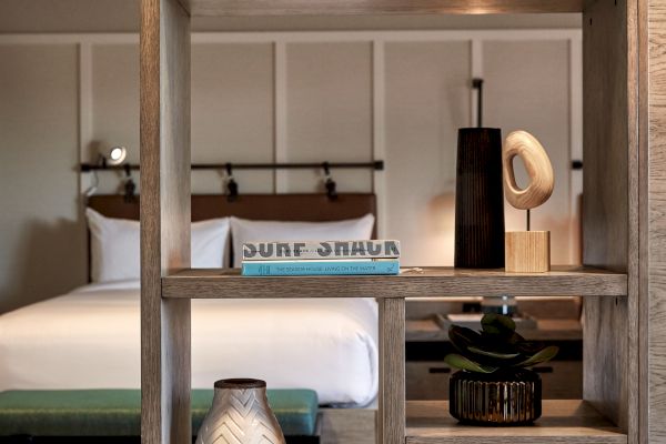 A modern room with a wooden shelf containing decorative items; in the background, there is a bed with white linens and pillows.