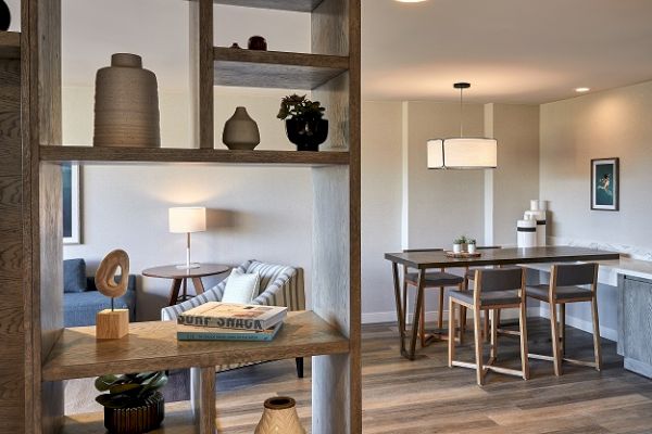 The image shows a modern living and dining area with wooden shelves, decorative items, a sofa, a lamp, a dining table, chairs, and wall art.