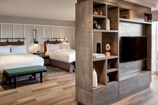 The image shows a modern hotel room with two double beds, a wooden partition with shelves and a TV, and a bench at the foot of one bed.