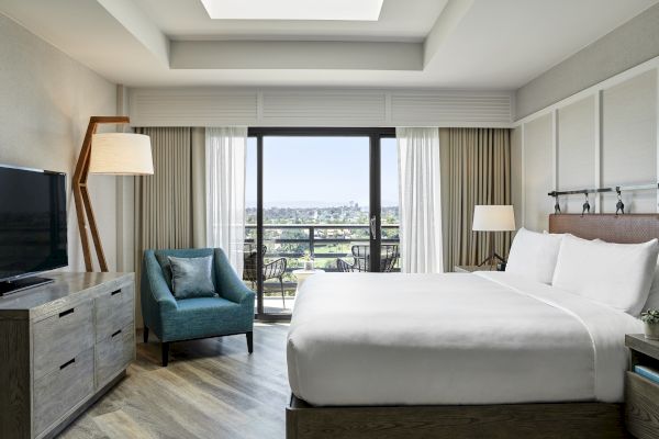 A well-lit hotel room with a king-sized bed, TV, armchair, dresser, floor lamp, and balcony doors opening to an outside view.