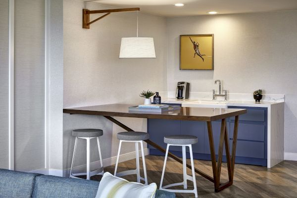 A modern kitchen setup with a wooden dining table, three stools, a sink, a coffee machine, a wall lamp, and a picture frame on the wall.