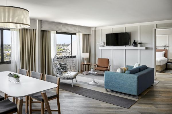 A modern living room with a dining area, sofa, chairs, wall-mounted TV, and hanging chair. Bedroom visible through the open door in the back.