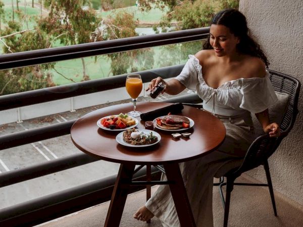 A person sits on a balcony enjoying a meal with various dishes and a drink on a round table. The view includes greenery and trees in the background.