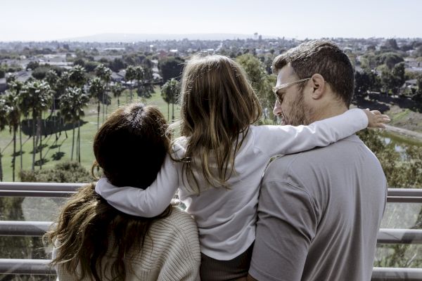 A family of three, viewed from behind, looks out at a scenic landscape from a balcony on a sunny day.