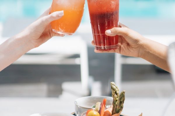 Two people clinking glasses with colorful drinks, with a spread of food and snacks in the foreground, near a poolside setting.