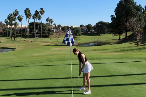 A person is playing golf, preparing to putt near a checkered flag on a sunny golf course with palm trees and a clear sky in the background.