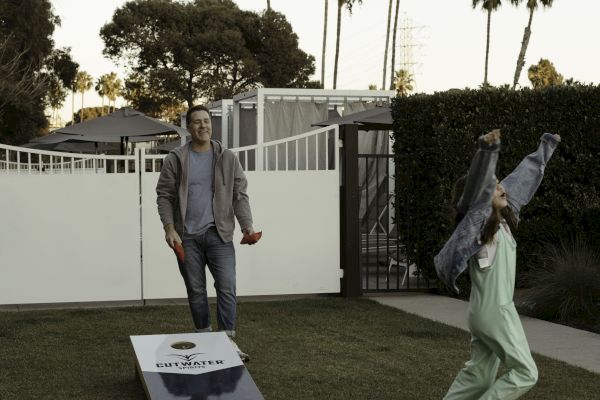 Two people playing cornhole in a backyard, with one person looking on and another celebrating. Palms and a gate are in the background.