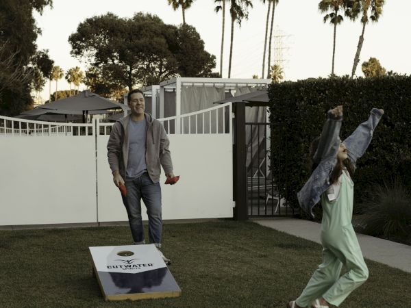 Two people playing cornhole in a backyard, with one person looking on and another celebrating. Palms and a gate are in the background.