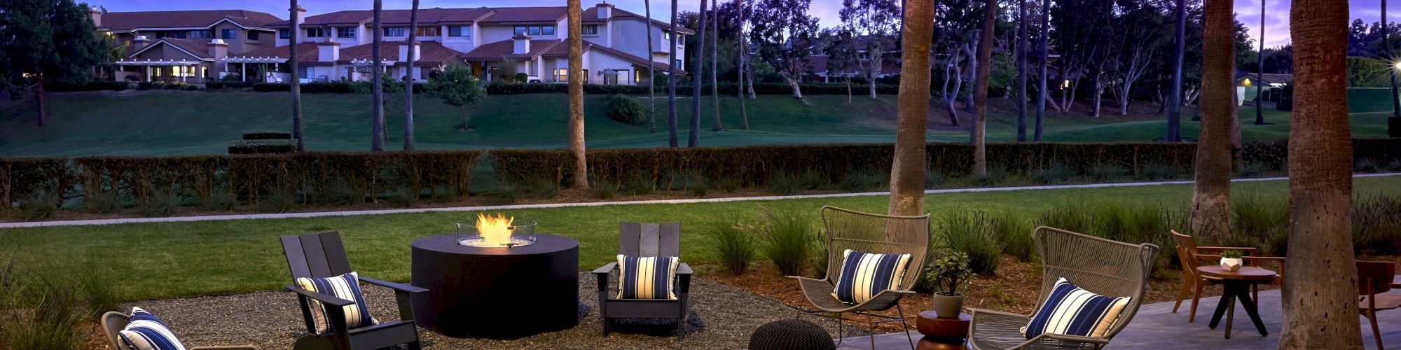 An inviting outdoor lounge area with seating, a fire pit, string lights, and palm trees, set against a twilight sky with surrounding buildings.