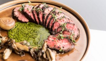 This image shows a plate of sliced beef garnished with herbs, served with a green sauce, mushrooms, and other garnishes on a brown plate.