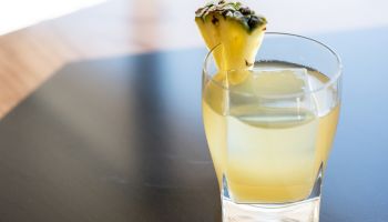 A glass filled with a yellowish drink, garnished with a slice of pineapple, is placed on a dark surface.