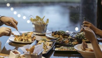 A dining table with various dishes, cutlery in use, and a view of a body of water and lights in the background, enhancing ambiance.