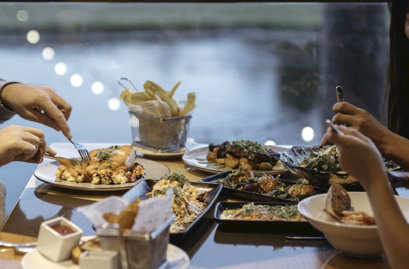 A dining table with various dishes, cutlery in use, and a view of a body of water and lights in the background, enhancing ambiance.