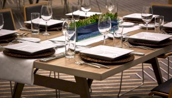 A neatly set dining table with plates, utensils, wine glasses, and a green centerpiece on a wooden table in a modern restaurant setting.