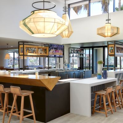 A modern bar setup with wooden stools, geometric lighting fixtures, and large windows. The bar has two sections with various bottles on display.