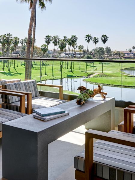 A modern patio with striped cushioned chairs, a sleek concrete table, overlooking a lush golf course with palm trees and a pond.