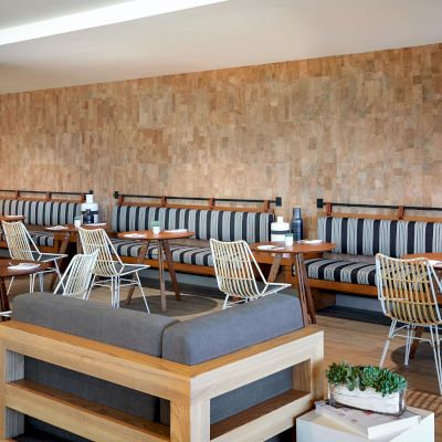 The image shows a modern, cozy seating area with striped benches, wooden tables, wire chairs, and potted plants against a textured wall background.