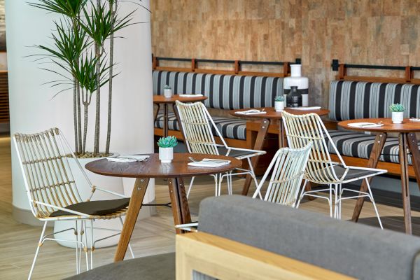 A modern café with tables, wire chairs, striped booth seating, plant décor, and a wooden wall. Empty seats and set tables await customers.