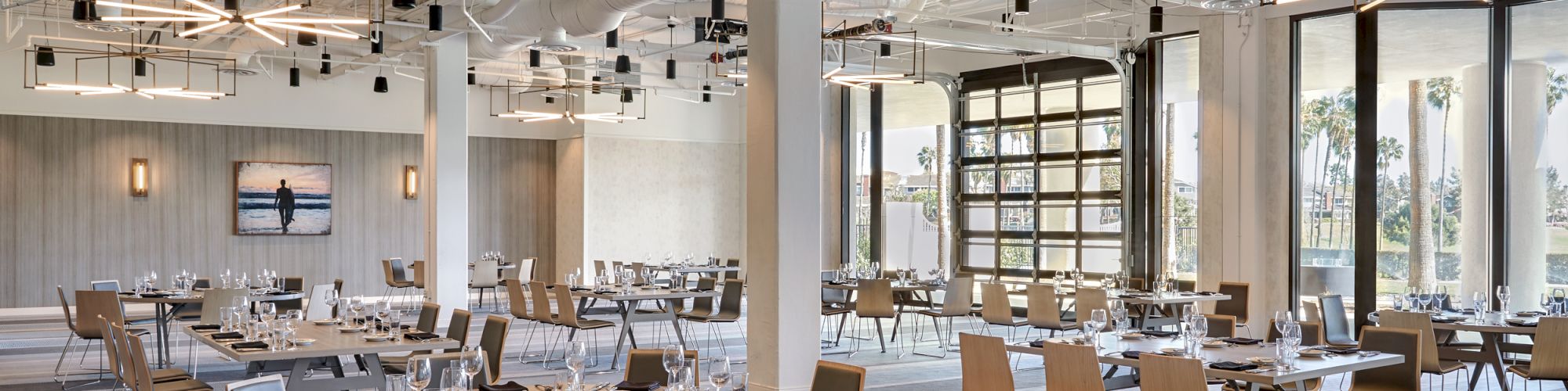 A modern dining area with wooden chairs, tables set with plates and glasses, and geometric ceiling lights in a spacious room with large windows.