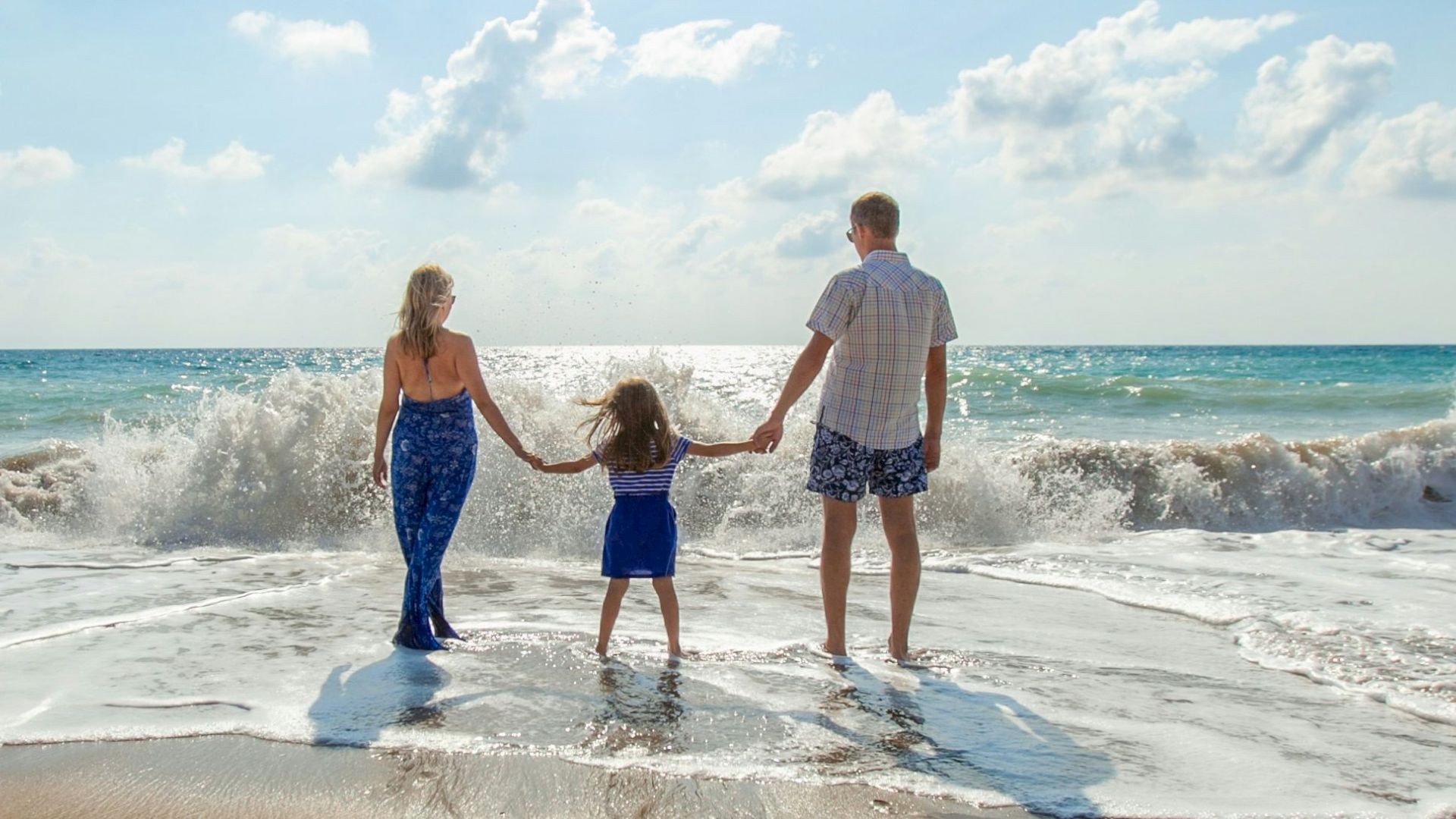 A family of three holds hands while standing at the edge of the ocean, facing the waves and horizon on a sunny beach day.
