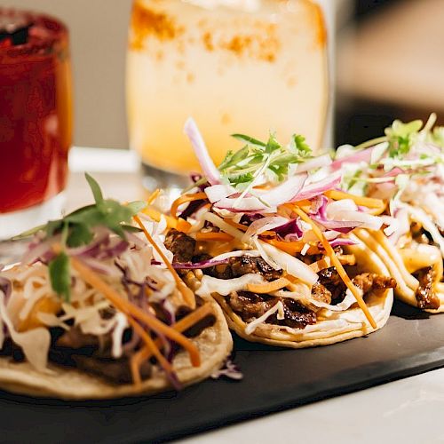 Three tacos topped with assorted vegetables sit on a black platter, accompanied by two colorful drinks in the background.