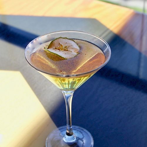 A martini glass filled with a light yellow cocktail, garnished with a thin slice of green apple, placed on a table with sunlight filtering through.