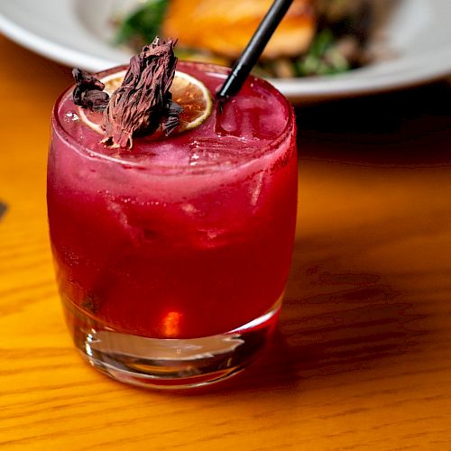 A vibrant red cocktail garnished with a dried slice of citrus and a black straw sitting on a wooden table, with a blurred dish in the background.