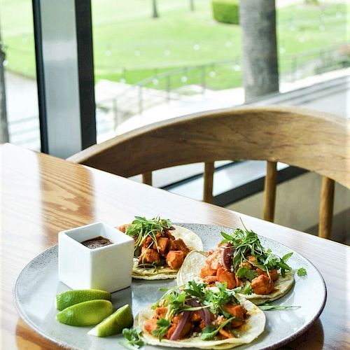 A plate with three tacos, fresh lime wedges, and a small container of sauce on a wooden table by a window with a green outdoor view.