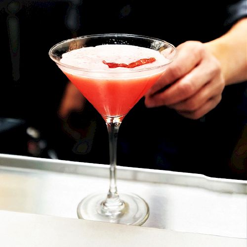 A hand holding a pink cocktail in a martini glass, garnished with a small slice of fruit.