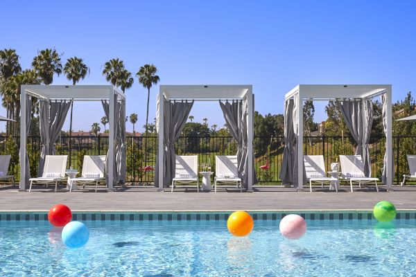 An outdoor pool with colorful beach balls floats in front of white cabanas and lounge chairs, with palm trees and blue skies in the background.