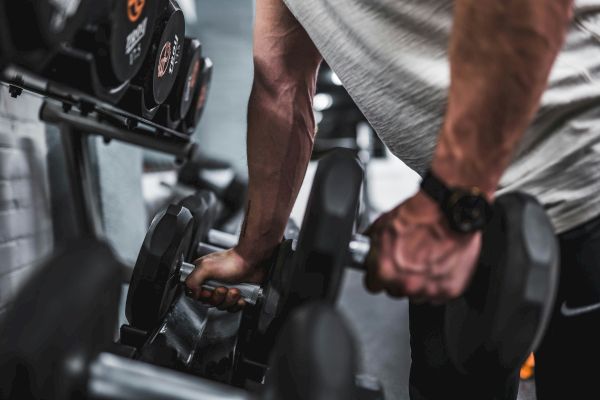 A person is picking up a dumbbell in a gym, with a focus on their arms and hands, and various weights are visible in the background.