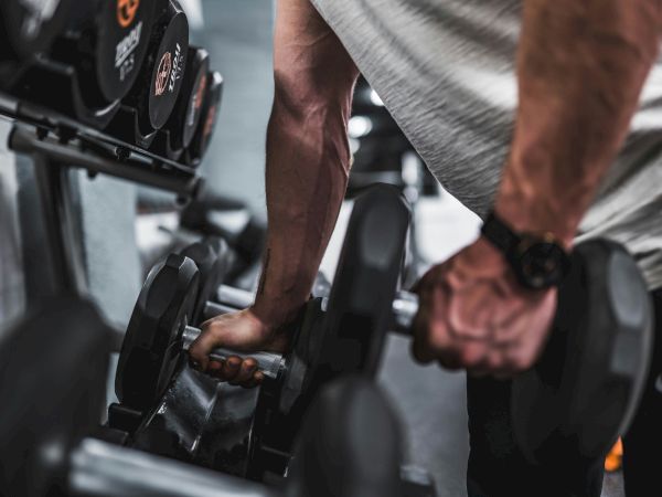 A person is picking up a dumbbell in a gym, with a focus on their arms and hands, and various weights are visible in the background.